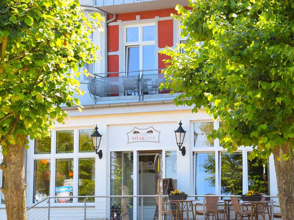 Terrasse des Alex Steakhouse mit Sitzplätzen unter Laubbäumen vor der weißen Außenfassade in Zinnowitz auf Usedom.