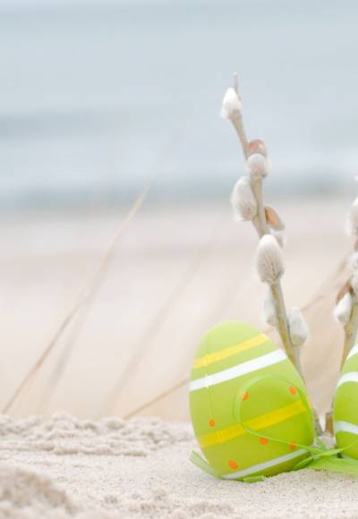 Drei bunt bemalte Ostereier und Weidenkätzchen liegen im Sand mit Blick auf die Ostsee.