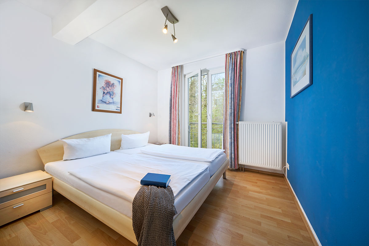 Helles Schlafzimmer der Ferienwohnung Strandperle mit Doppelbett, blauer Wand und Balkon.