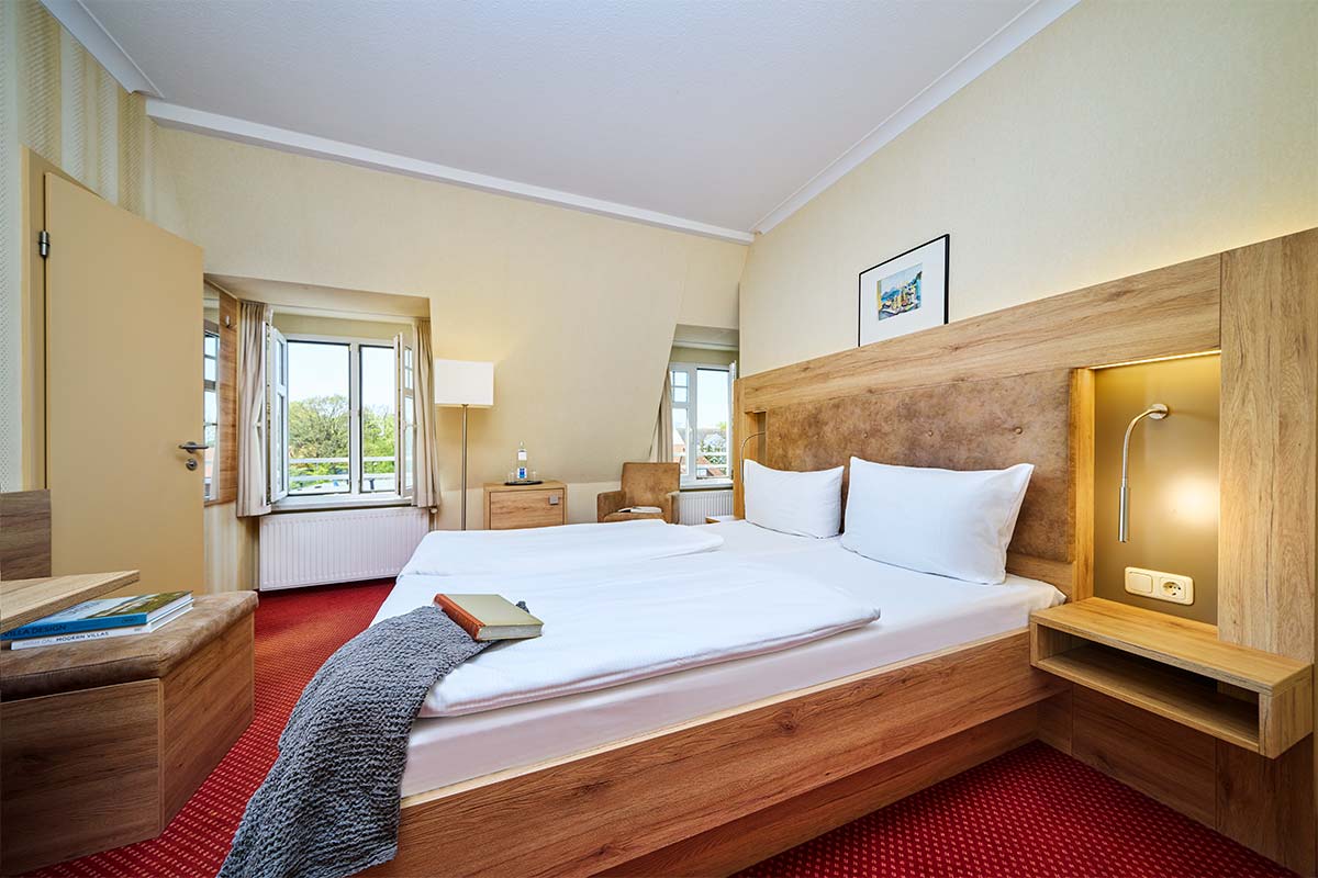Doppelzimmer mit Bett, Nachttischlampen, Sessel, Schreibtisch und Fenster mit Blick zur Ostsee im Hotel Kormoran.