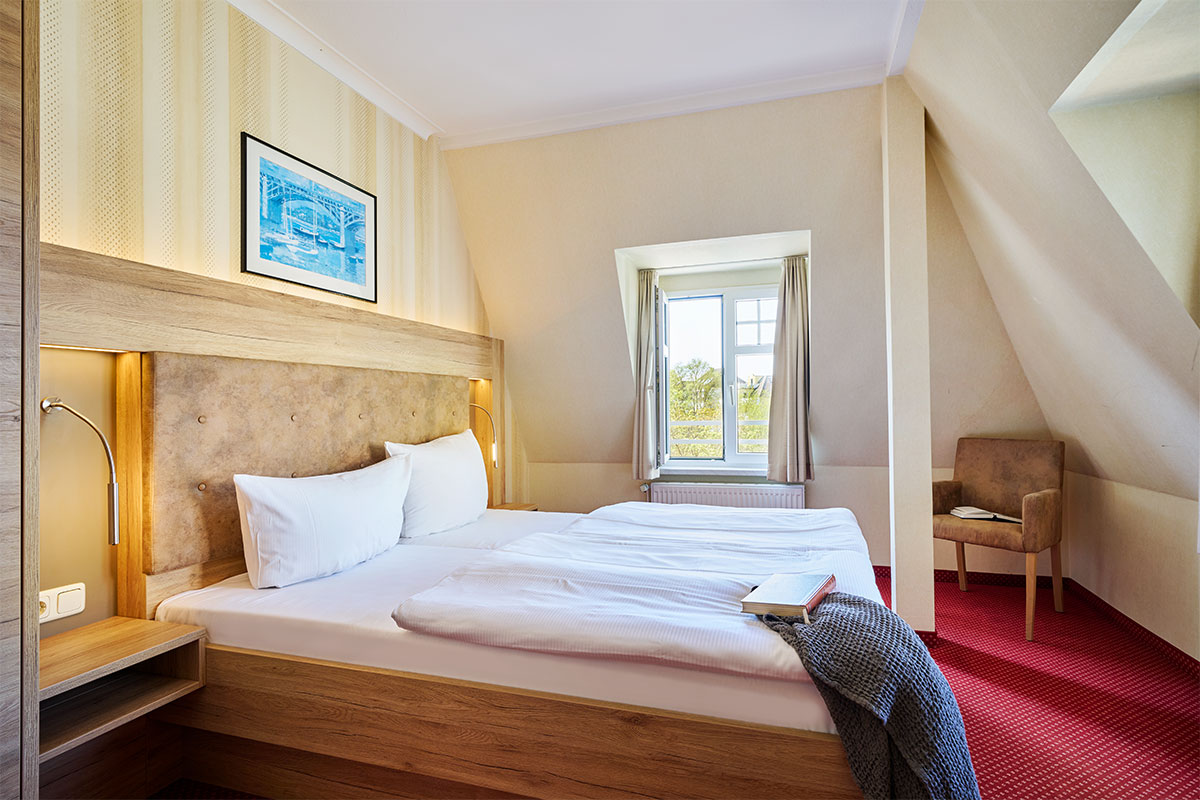 Großes Doppelzimmer mit getrenntem Schlafbereich, Fenster, Bett, Sessel und rotem Teppich im Hotel garni Kormoran.