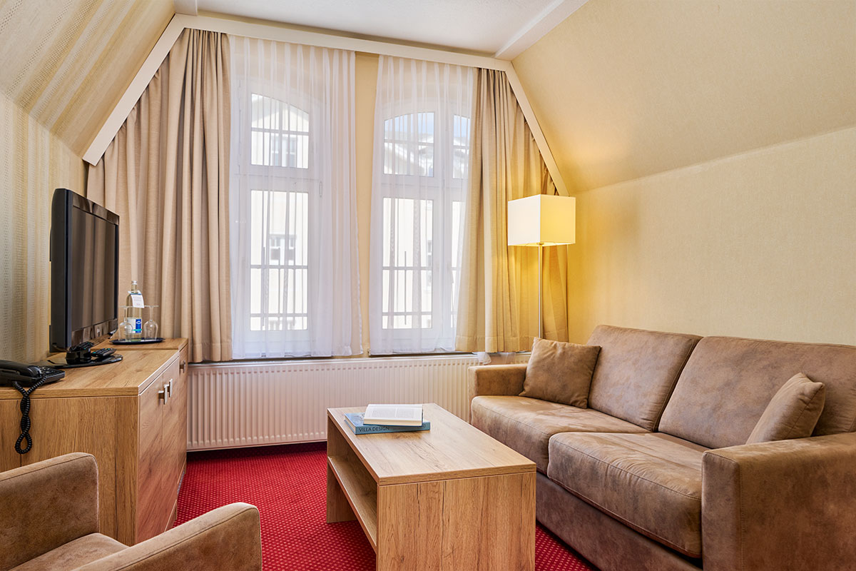 Wohnbereich mit Sofa, Sessel, TV, Vorhängen und Couchtisch im großen Doppelzimmer des Hotels Kormoran an der Ostsee.