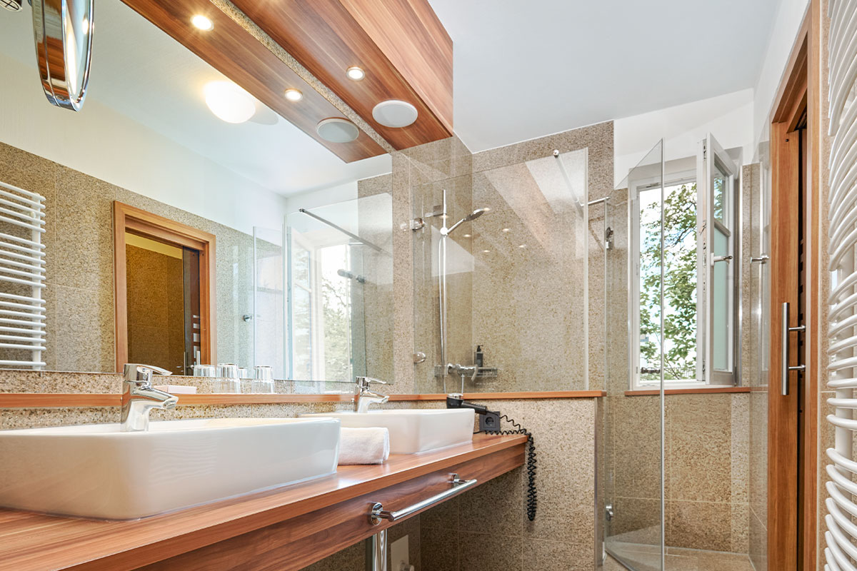 Badezimmer mit zwei Waschbecken, Glasdusche, Fenster und Spiegel im Strandhotel Kormoran.