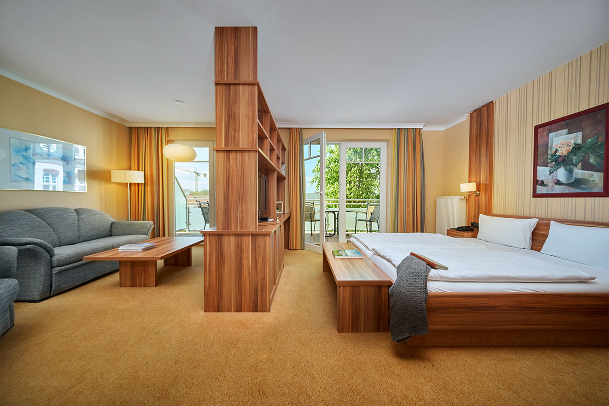 Großzügiges Zimmer im Strandhotel Kormoran mit Balkon zur Landseite, Doppelbett, Sofa, TV und Tisch.