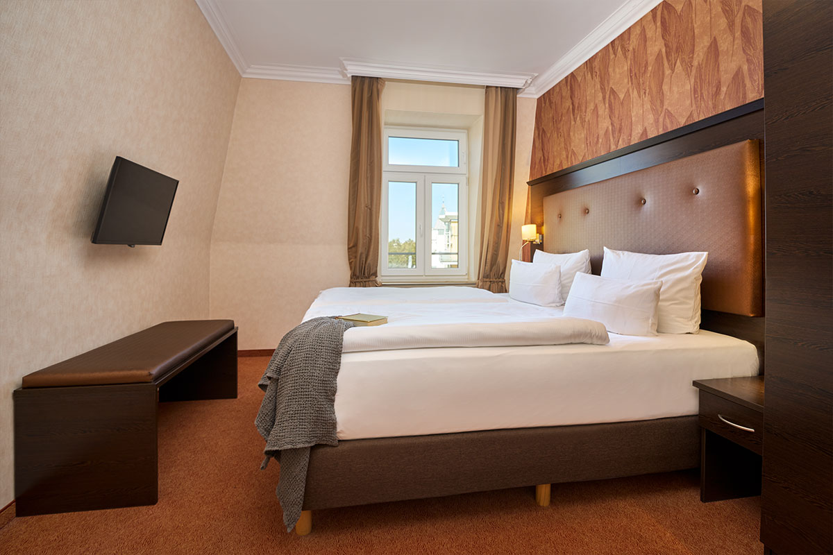Modernes Doppelzimmer im Strandhotel Preußenhof mit Fenster, TV, Sitzbank und gemütlichem Bett.