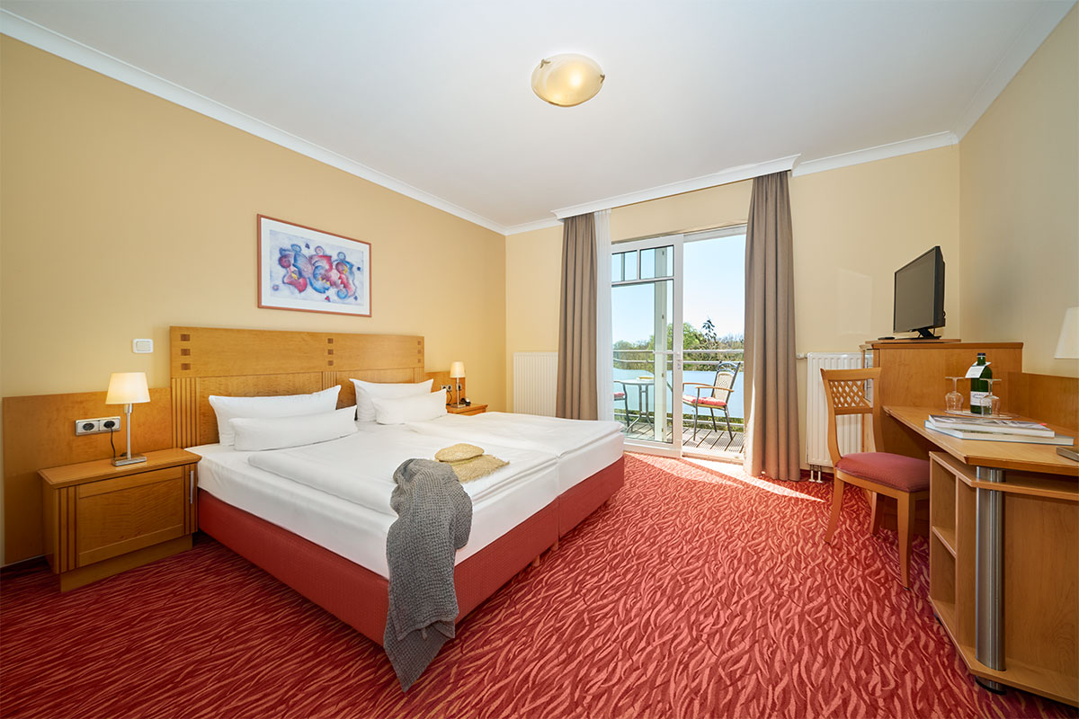 Porträt eines Doppelzimmers mit Schreibtisch, Bett, Balkonmöbeln und Ausblick ins Grüne im Strandhotel Prinz von Preussen.