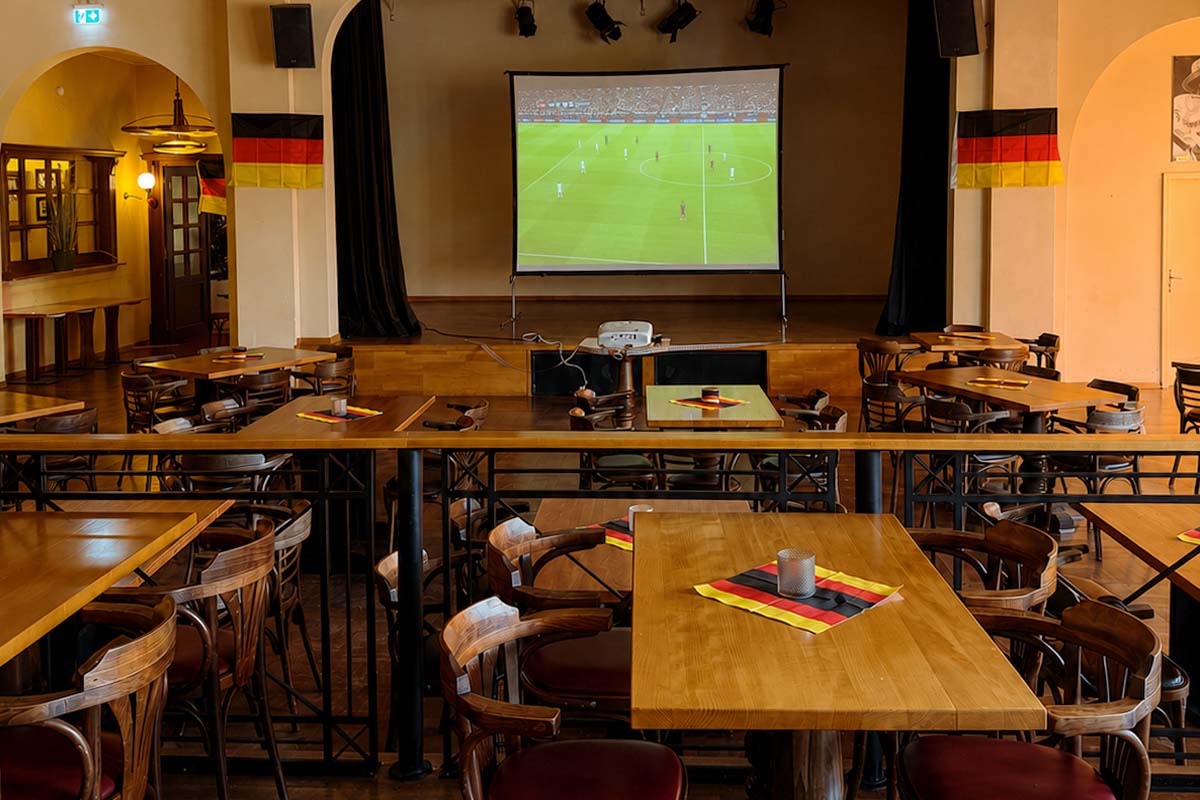 Festsaal Belle Époque mit Tischen und Stühlen aus Holz, Deutschland-Servietten auf den Tischen und einer Leinwand mit einem Fußballspiel im Hintergrund