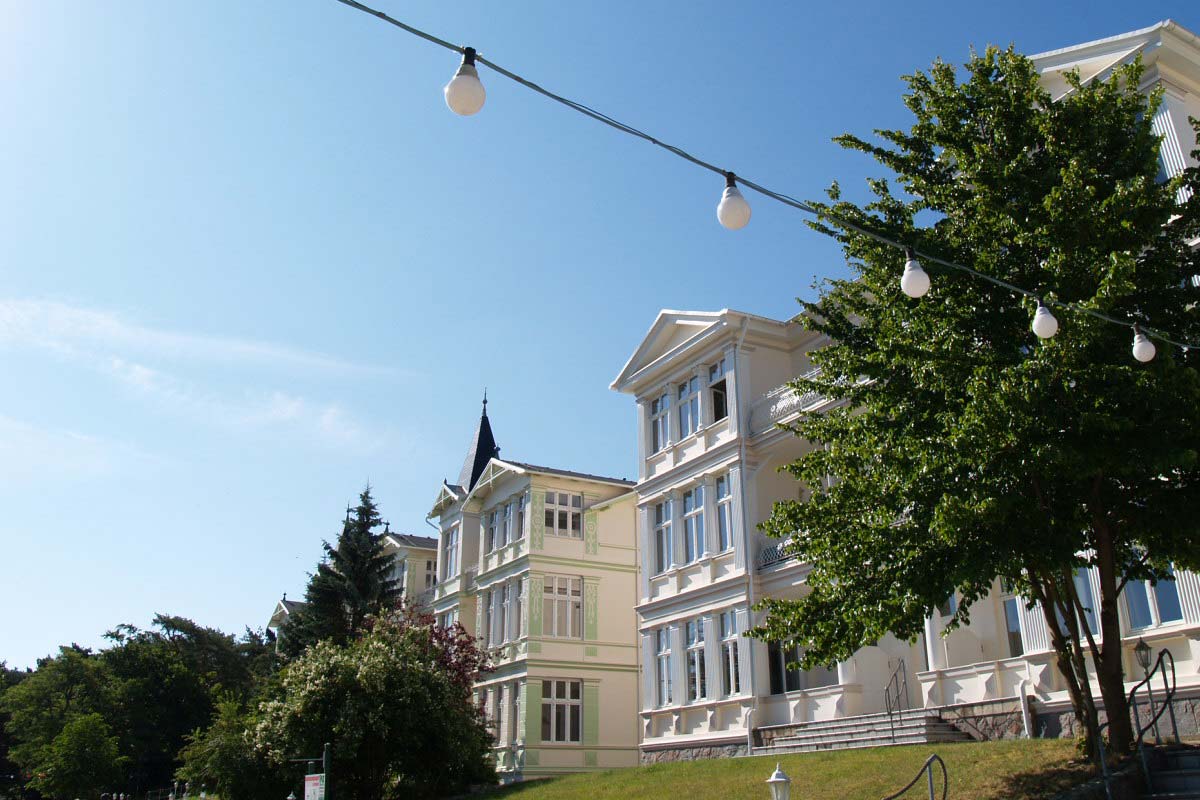 Außenansicht vom Zinnowitzer Hof von Schöner Inseln auf Usedom mit heller Fassade, Balkonen und Bäumen im Vordergrund.