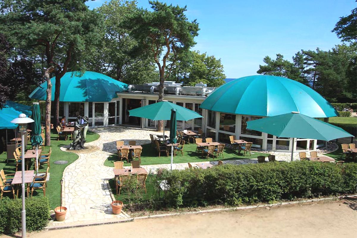Sommerliches Promenadenrestaurant von Schöner Inseln mit türkisfarbenen Kuppeln und Sitzplätzen unter Pinien am Meer.